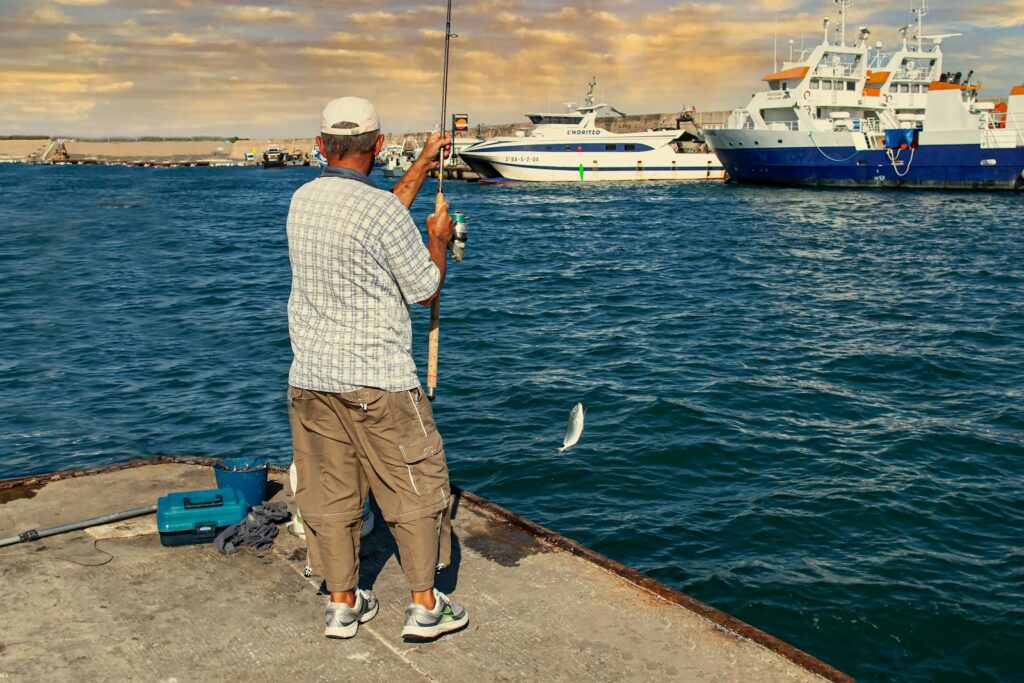 A fisherman reels in a catch by the pier at L'Ametlla de Mar, Spain with boats in the background.