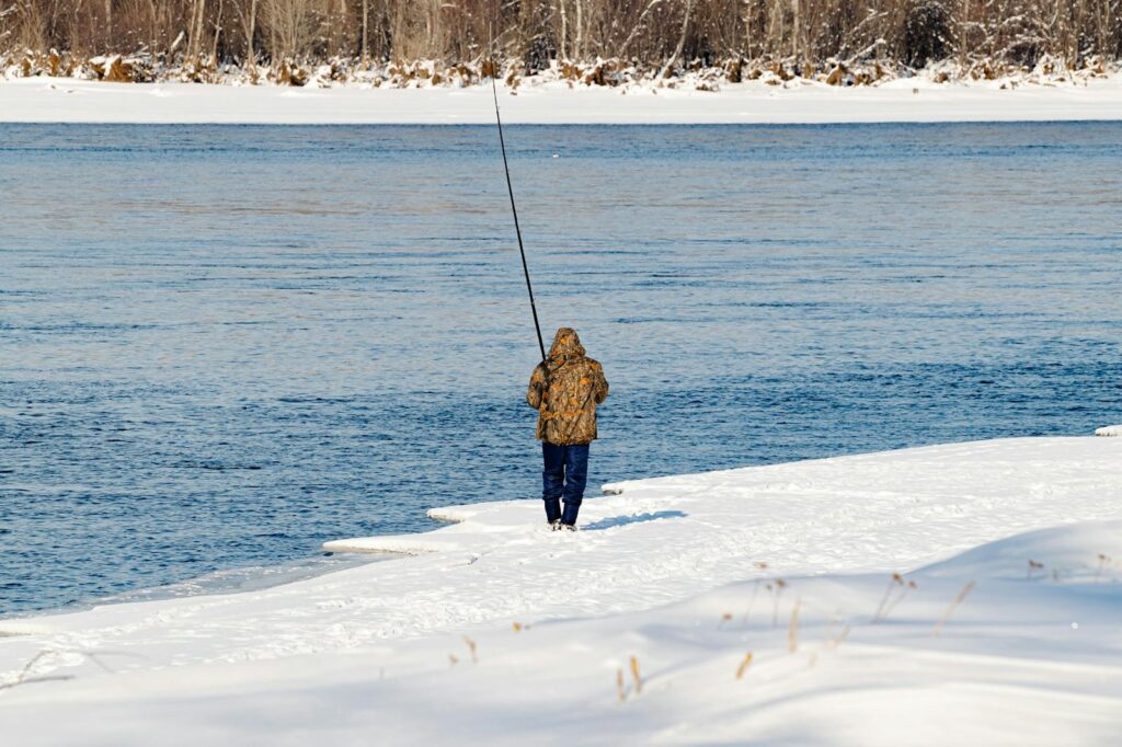 A person dressed warmly fishes alone by a snow-covered river in a winter landscape.