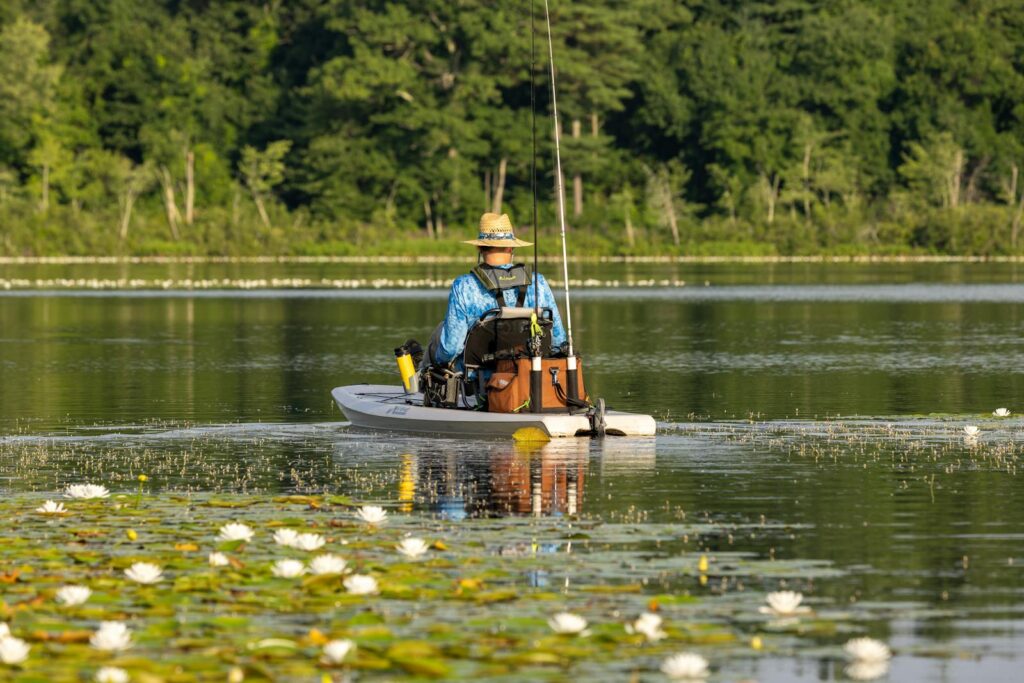 A man in a hat fishes from a kayak on a tranquil lake surrounded by lily pads and lush greenery.