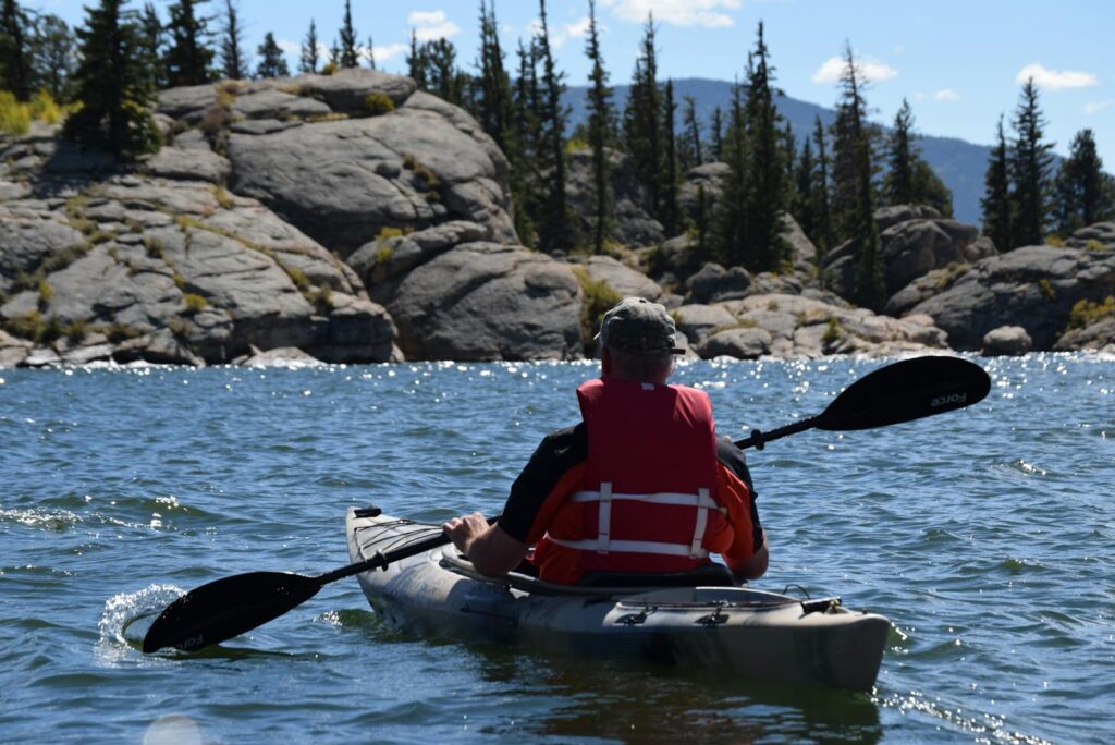 Person kayaking on a mountain lake surrounded by rocks and trees under a clear blue sky.