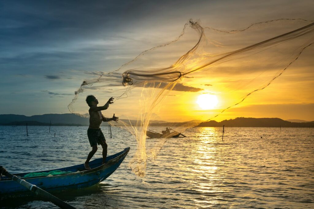 A fisherman skillfully casts a net at sunrise, silhouetted against a golden sky over a calm lake.