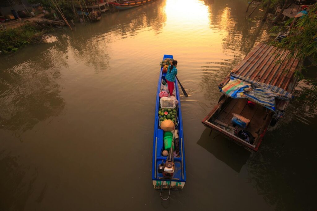 A woman rows a boat laden with fruits on a serene river during sunset, capturing the tranquility of the moment.