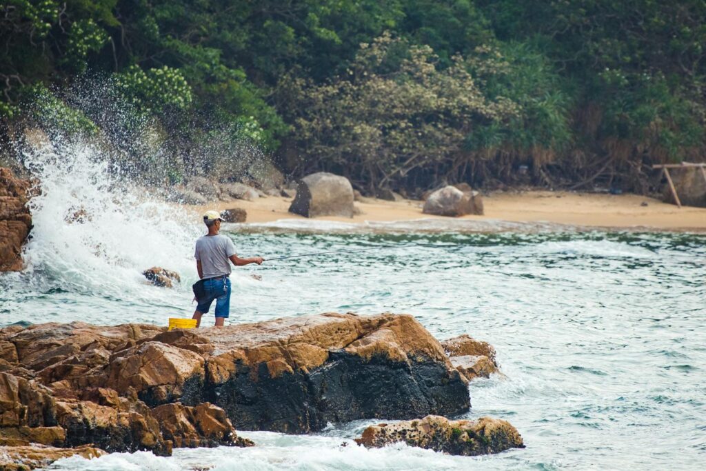 A man fishing on a rocky shore by the ocean in Hong Kong, with waves crashing nearby.