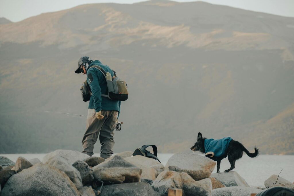 A man and his dog fishing on a rocky lakeshore, surrounded by mountains.