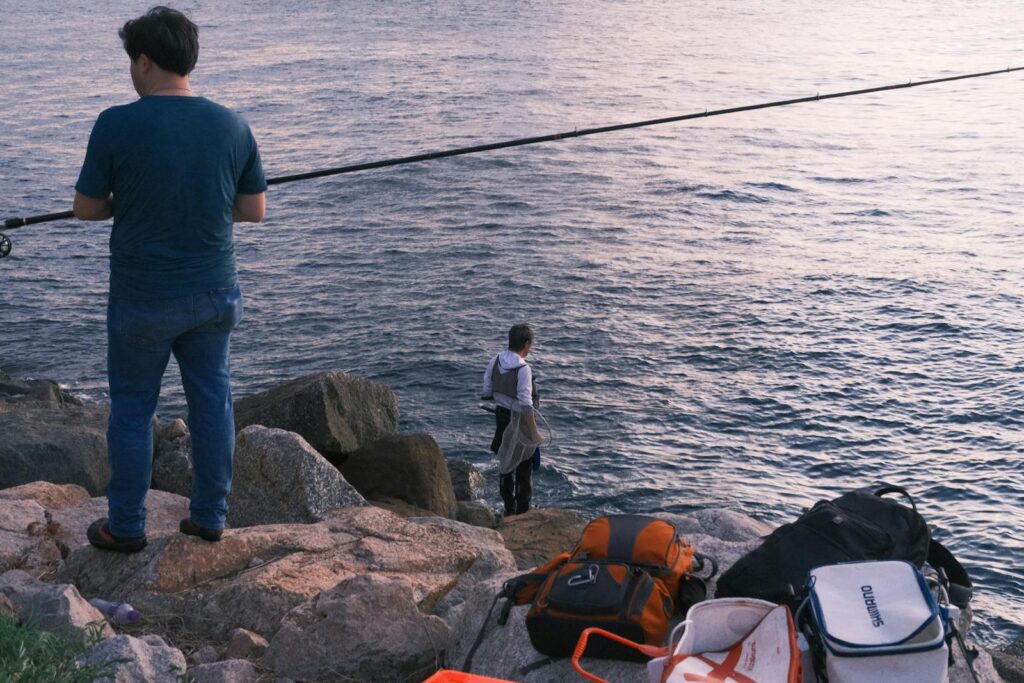 Two men are fishing from rocky shorelines at sunrise, capturing peaceful coastal scenes.