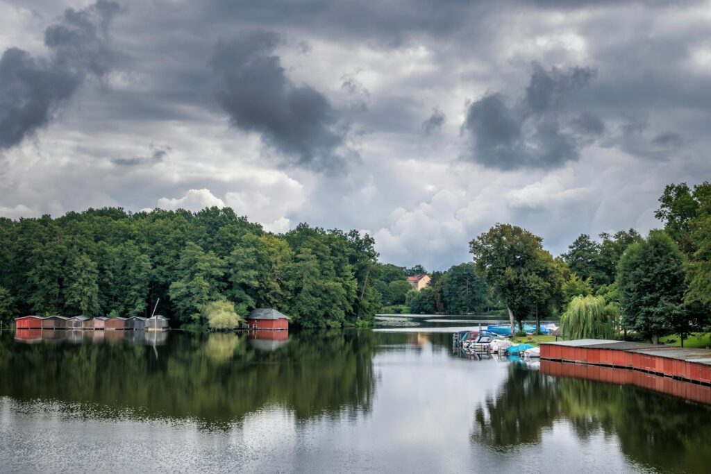 Serene lakeside scene featuring boats, a dense forest, and overcast sky reflecting on the water.