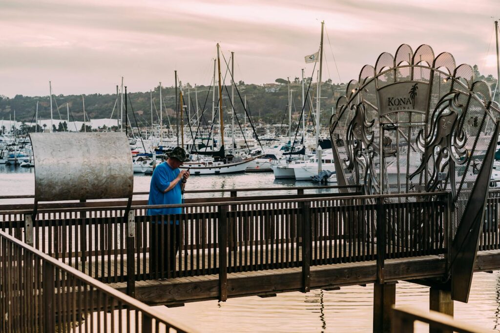 A man stands on a wooden dock at Kona Kai Marina, with yachts and harbor in the background.