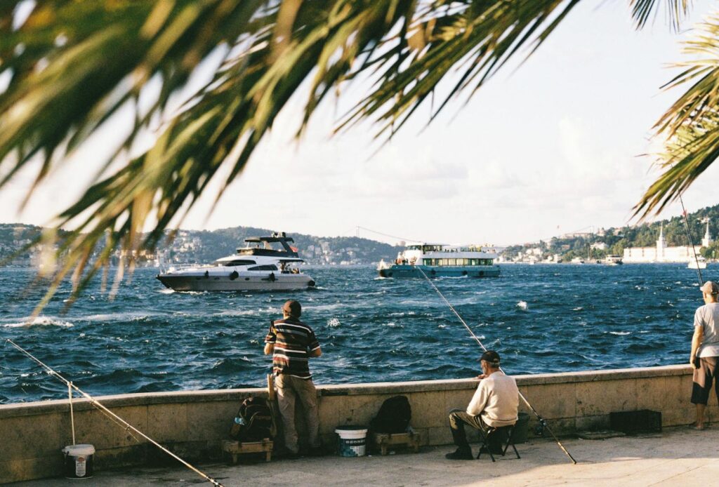 Fishermen fishing by a vibrant waterside with boats and scenic backdrop. Perfect serene day outdoors.