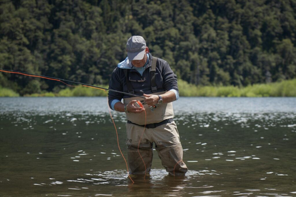 Man fly fishing in Patagonia's tranquil lake with lush forest backdrop.