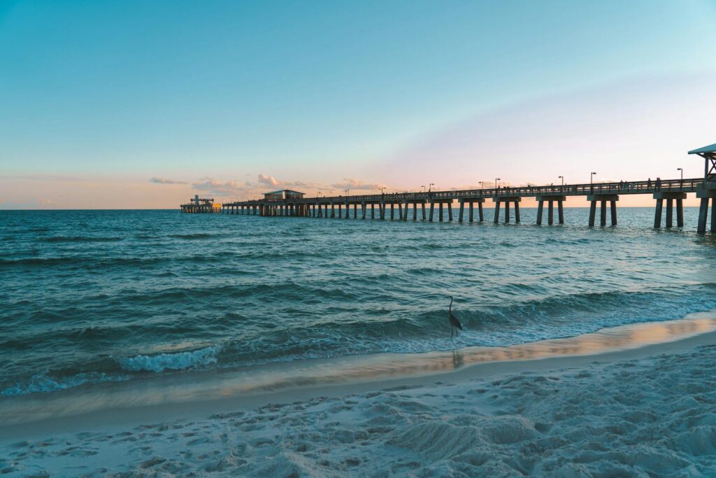 A serene view of a long pier extending over the ocean at sunset, with gentle waves on the sandy shore.