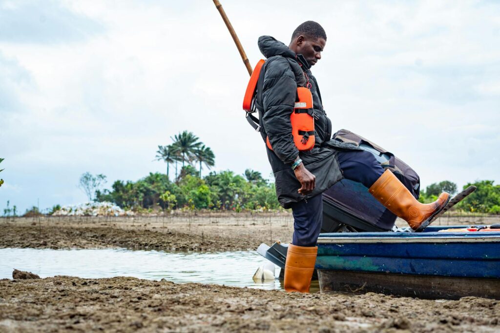 A fisherman in waterproof boots and life jacket stepping from a boat onto a muddy shore.
