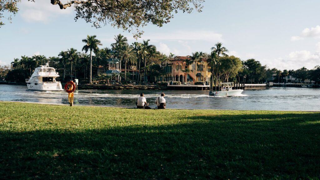 A peaceful riverside view featuring palm trees, boats, and people relaxing by the water.