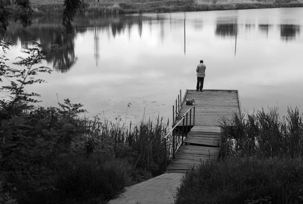 Black and white image of a man fishing on a pier by a calm lake.