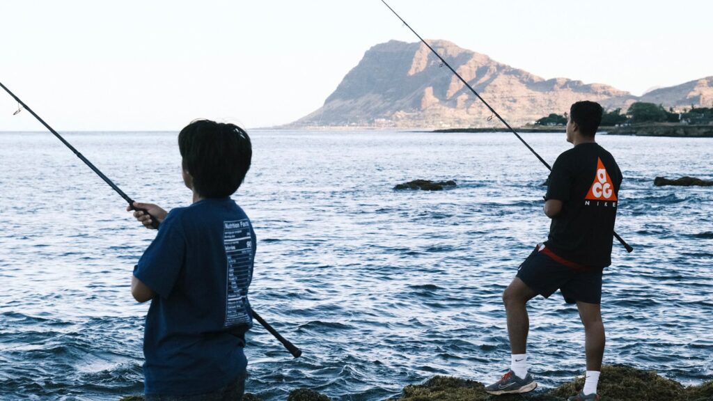 Two people fish by the sea, holding rods against a backdrop of calm water and distant mountains. The scene feels peaceful and relaxed.