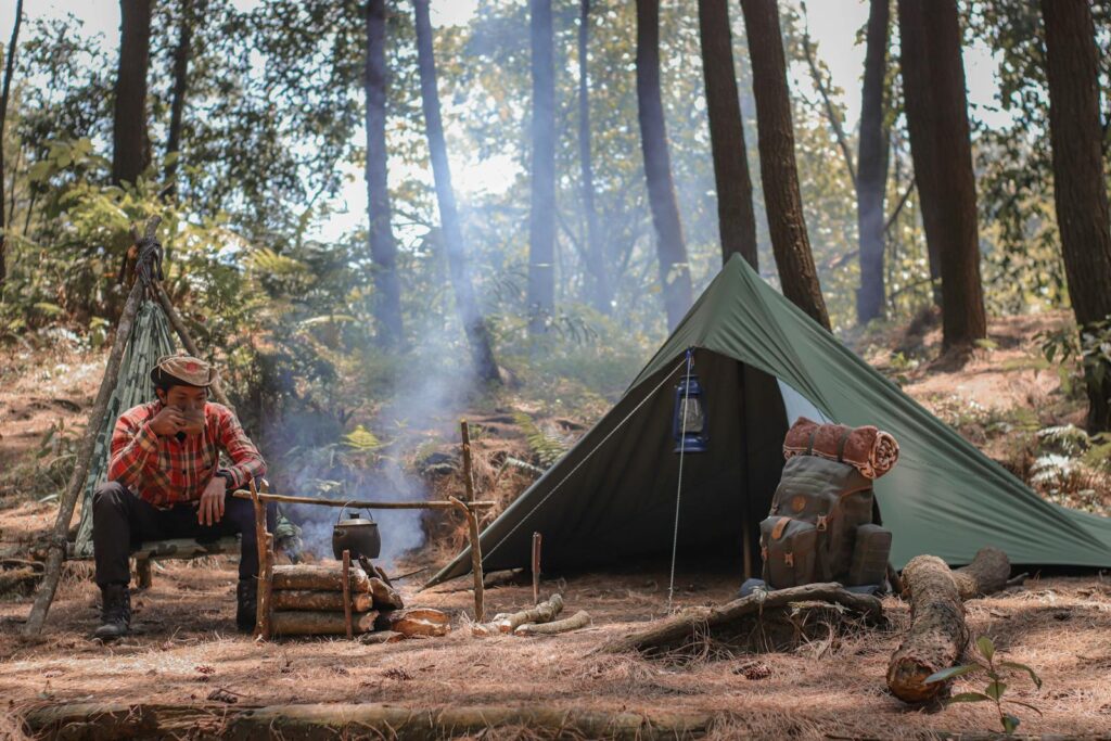 Man camping in forest, sitting by campfire with tent and backpack.