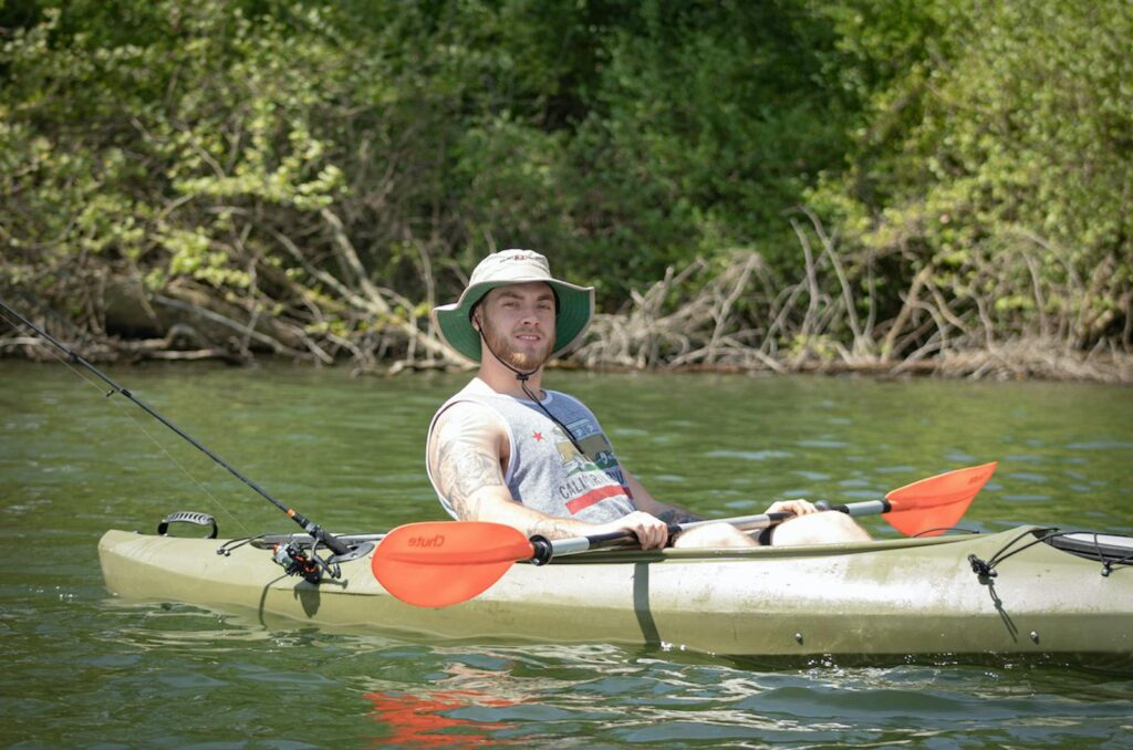 A man in a hat enjoys a peaceful day kayaking and fishing on a sunny river.
