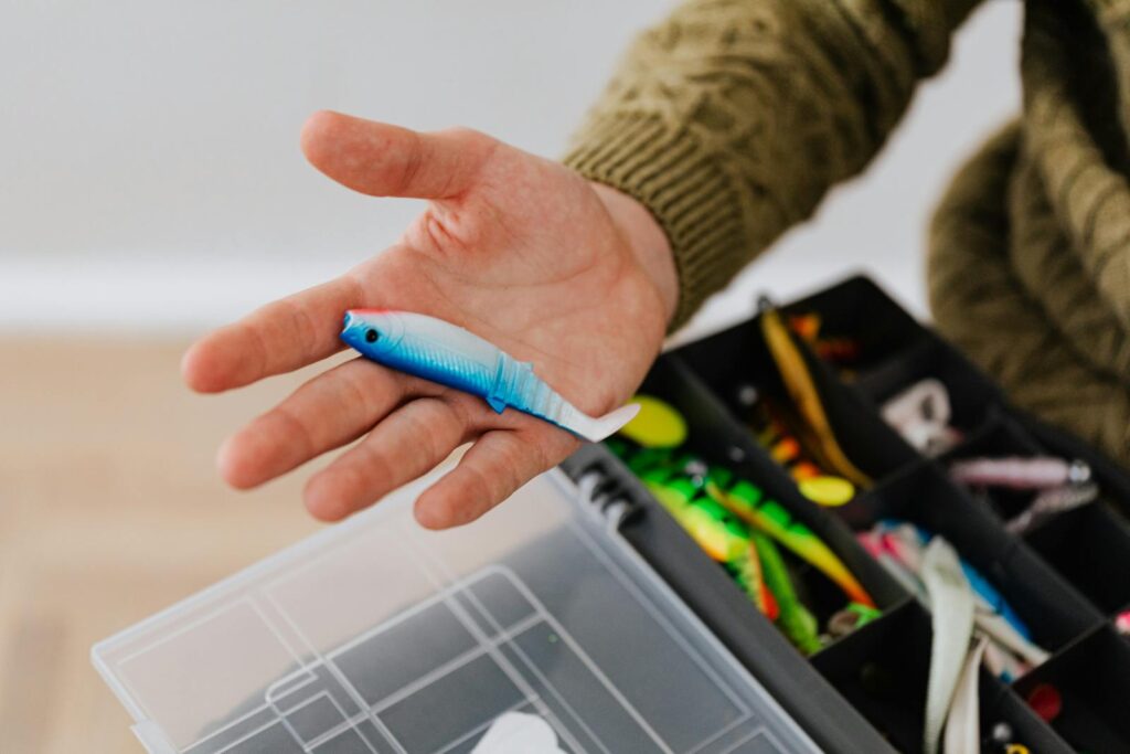 Close-up of a hand holding a fishing lure over a tackle box indoors.