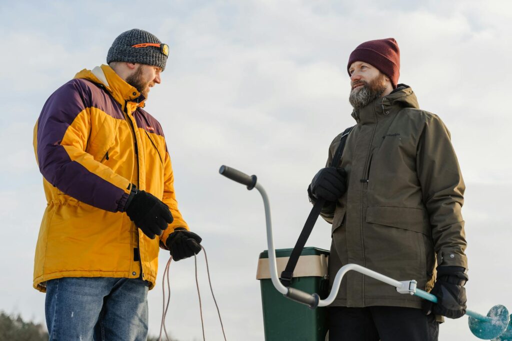 Two bearded men in winter clothes ice fishing outdoors with equipment.