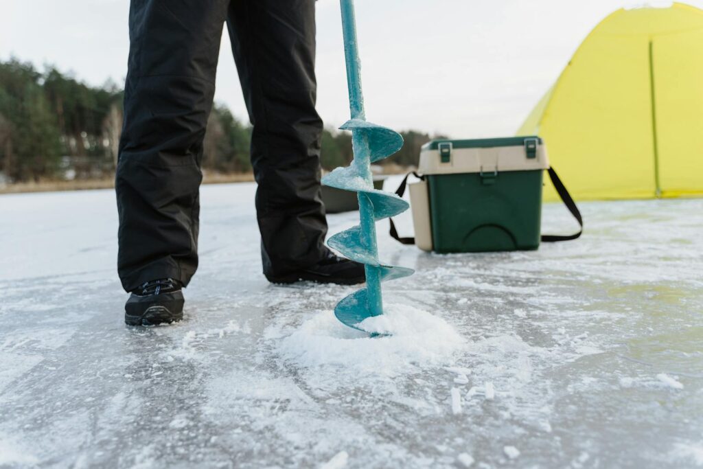 Person setting up ice drill for fishing on a frozen lake with equipment nearby.