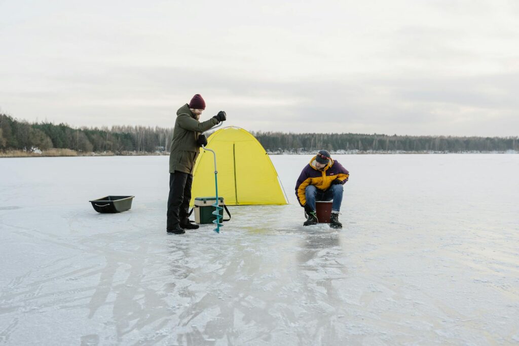 Two men ice fishing near a yellow tent on a frozen lake in winter.