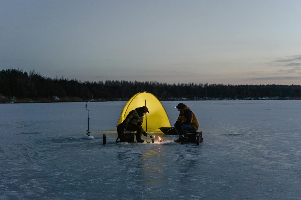 Two people camp on a frozen lake, cooking by a tent at twilight.
