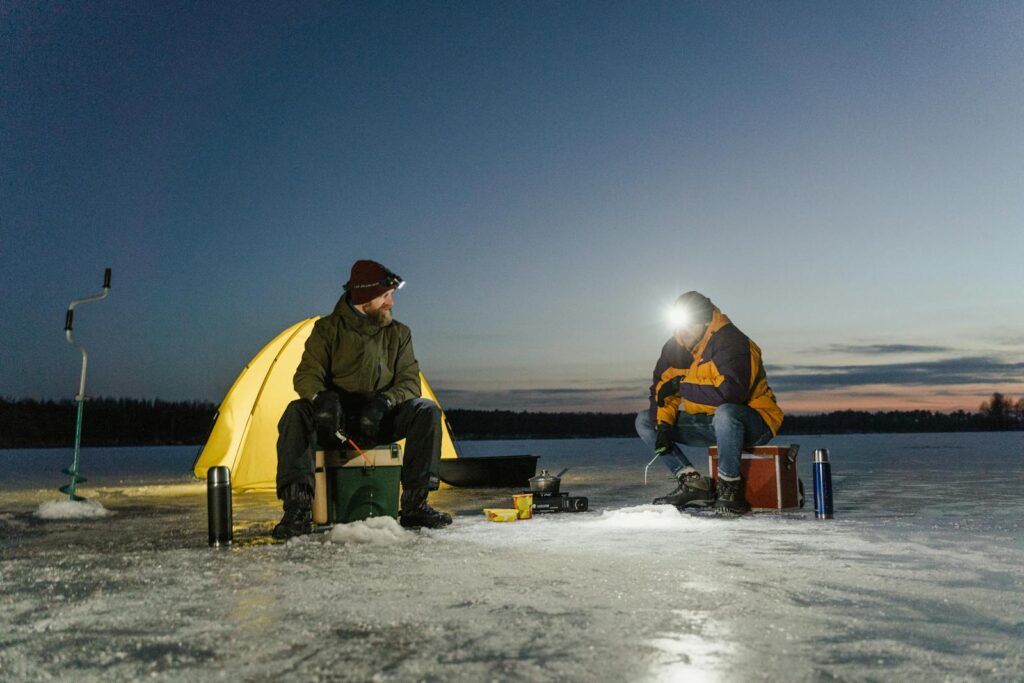 Two men ice fishing by a tent during winter dusk. Adventure on a frozen lake.