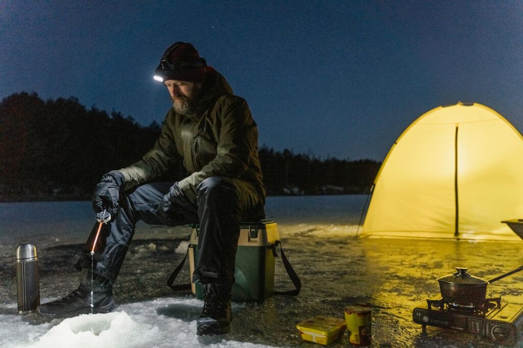 A man ice fishing at night next to a glowing tent on a frozen lake.