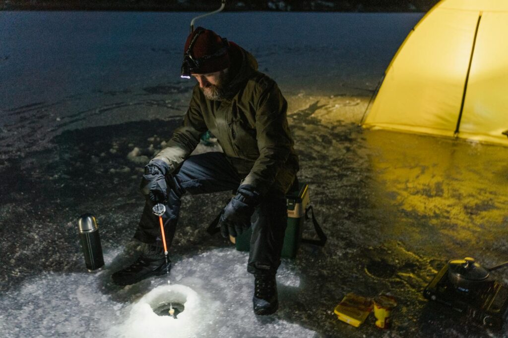 A lone fisherman enjoys ice fishing on a frozen lake at night, illuminated by a camping light.