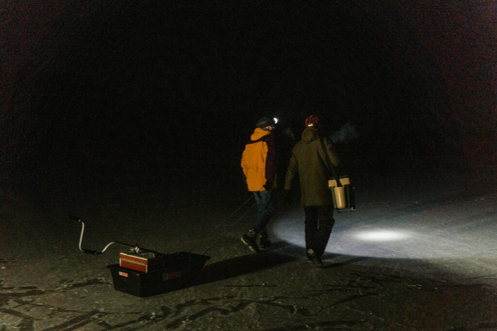 Two men ice fishing at night, walking on a frozen lake with headlamps and pulling gear.