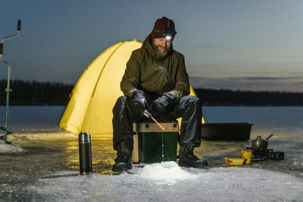 A man ice fishing by tent on a frozen lake during winter night, capturing outdoor adventure.
