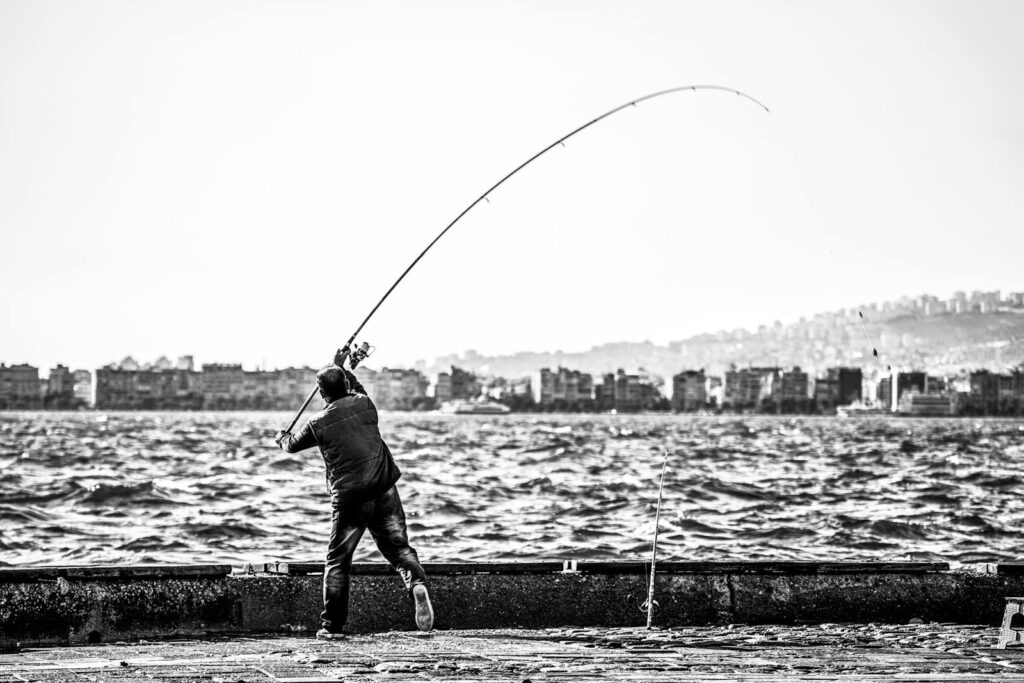 A man casting a fishing line by the sea with a cityscape in the background.