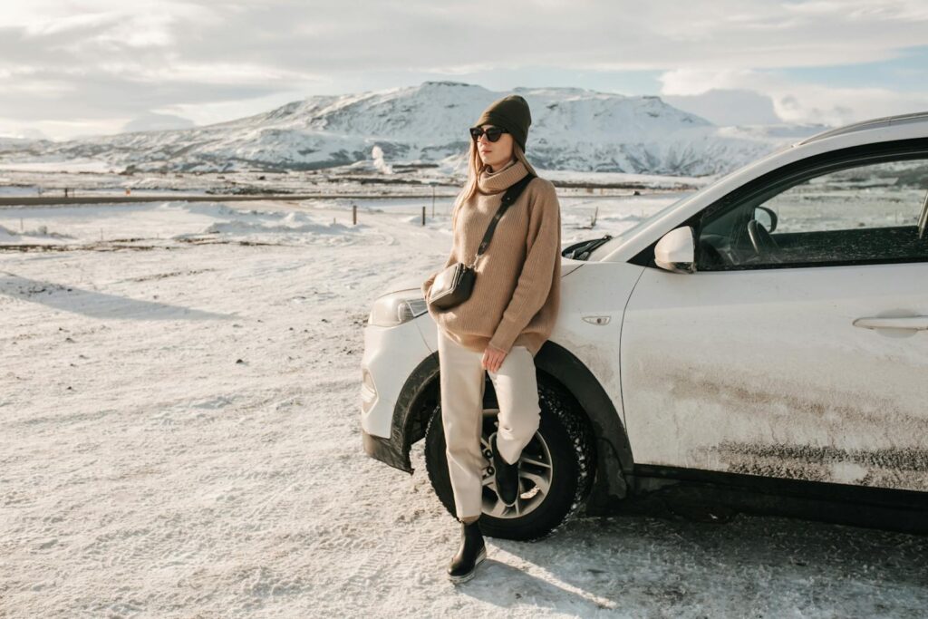 A fashionable woman poses by a vehicle in a snowy Icelandic landscape with mountains in the background.