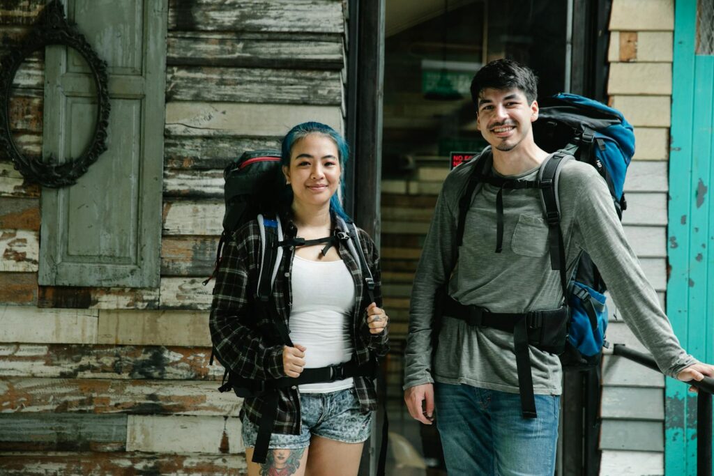 Young smiling diverse couple of travelers in casual clothes and with backpacks standing together near wooden wall in light room and looking at camera