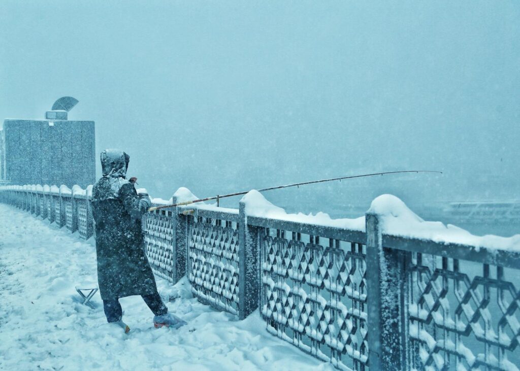 Person fishing on a snow-covered bridge during a heavy winter snowstorm