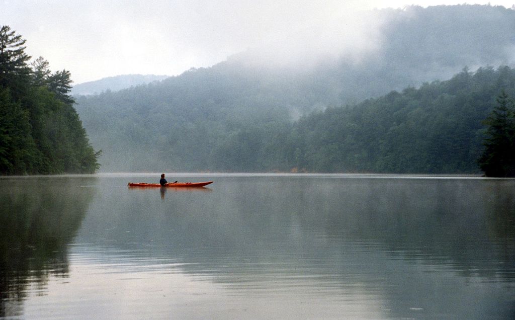 A lone kayaker in an orange kayak paddles on a serene lake surrounded by misty, forested hills, creating a peaceful and tranquil scene.