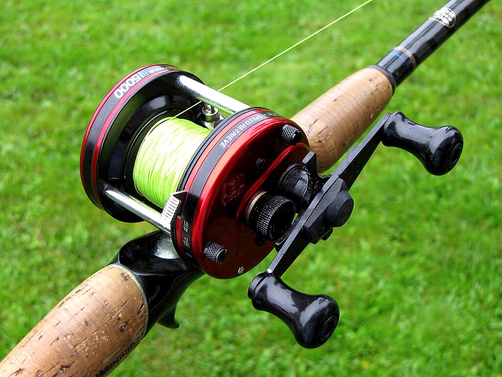 Close-up of a red fishing reel with bright green line, attached to a cork-handled rod, set against a grassy background, conveying readiness for fishing.