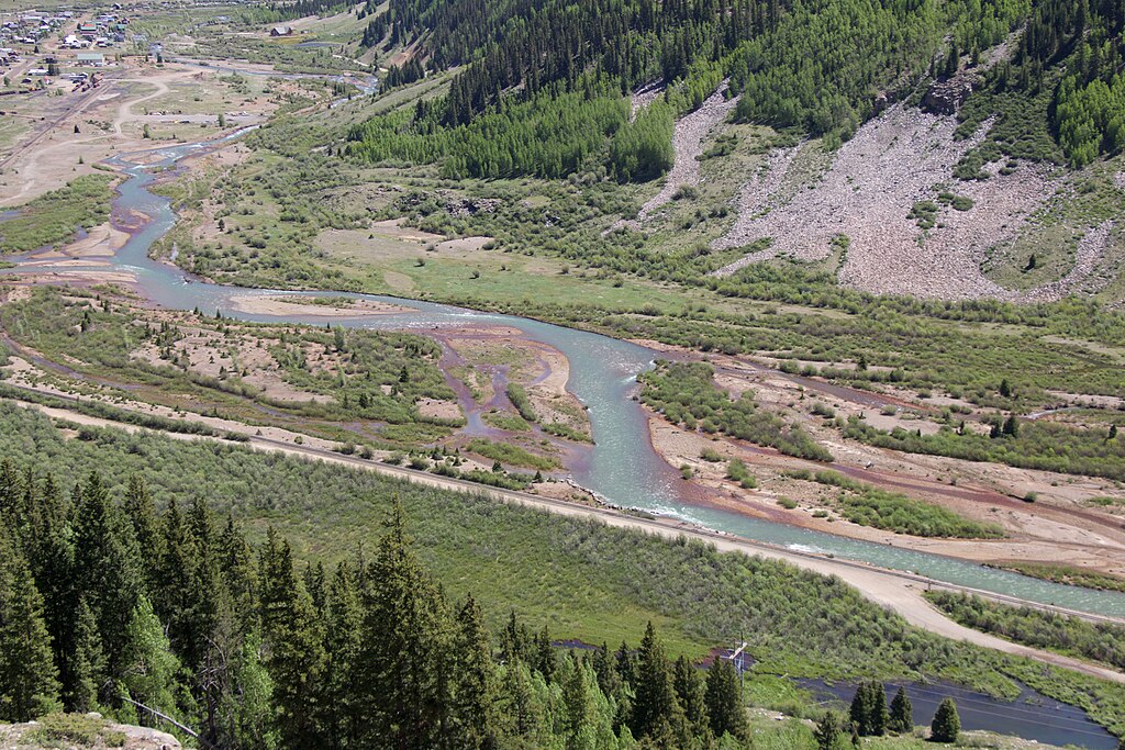 A meandering river flows through a lush green valley with dense forests and rocky hillsides. A small town is visible in the distance, conveying tranquility.