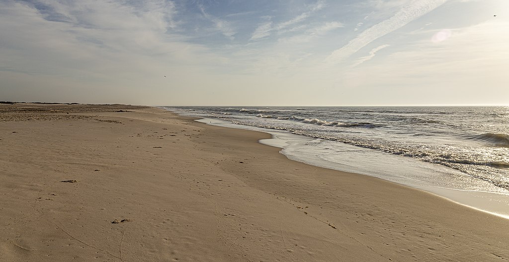 Wide sandy beach with gentle waves on a serene, sunny day. The sky is partly cloudy, and the horizon stretches far into the distance, conveying calm.