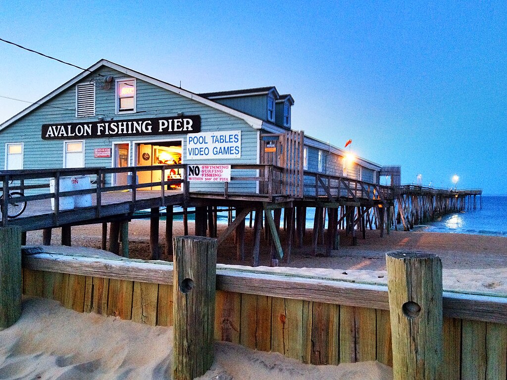 Wooden pier extending over the ocean at dusk with a brightly lit building labeled "Avalon Fishing Pier." Signs advertise pool tables and video games.