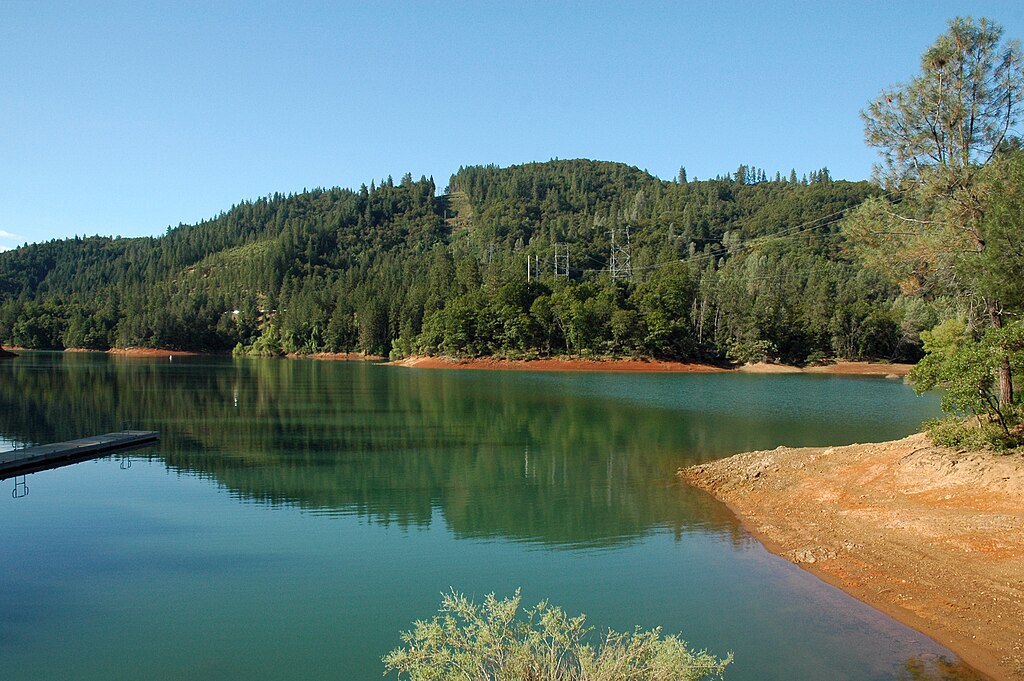 A calm lake reflecting a forested hillside under a clear blue sky. A small dock protrudes into the water, creating a serene, peaceful atmosphere.