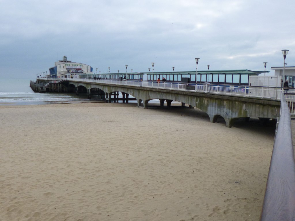 A long pier extends over sandy beach towards a calm sea under a cloudy sky. Lamps line the pier, leading to a building marked "PIER THEATRE."