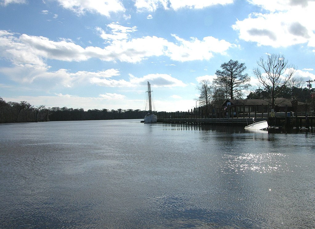 Calm river scene with a sunken boat near a wooden dock on the right, surrounded by bare trees and buildings under a partly cloudy blue sky.