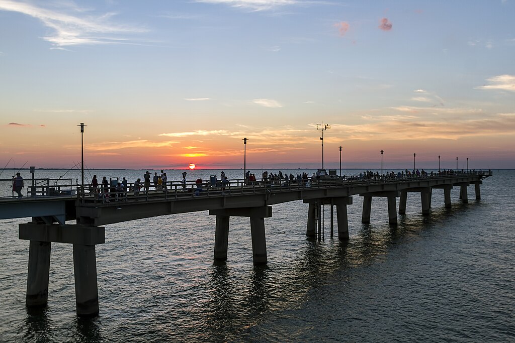 A long pier with silhouetted people extends over calm water, leading to a vibrant sunset on the horizon, creating a tranquil and serene atmosphere.