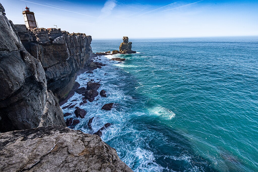 Rocky cliffside with a lighthouse stands above the crashing blue sea. The sky is clear, adding a sense of tranquility and vastness to the scene.