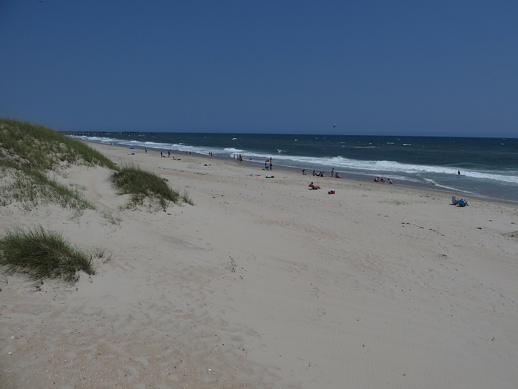 Sandy beach with scattered grass dunes, people relaxing near the shoreline, and a distant pier under a clear blue sky, capturing a peaceful coastal scene.