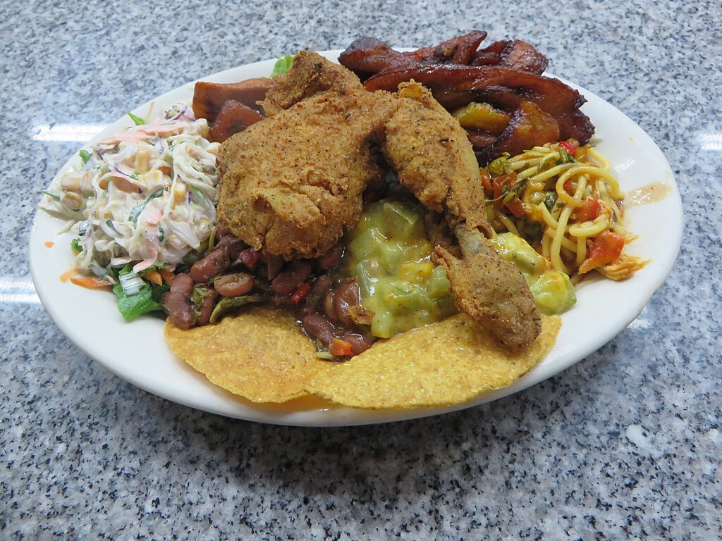 A plate with fried chicken, crispy plantains, spaghetti, cole slaw, beans, avocado, and tortillas on a speckled gray countertop, conveying a hearty meal.