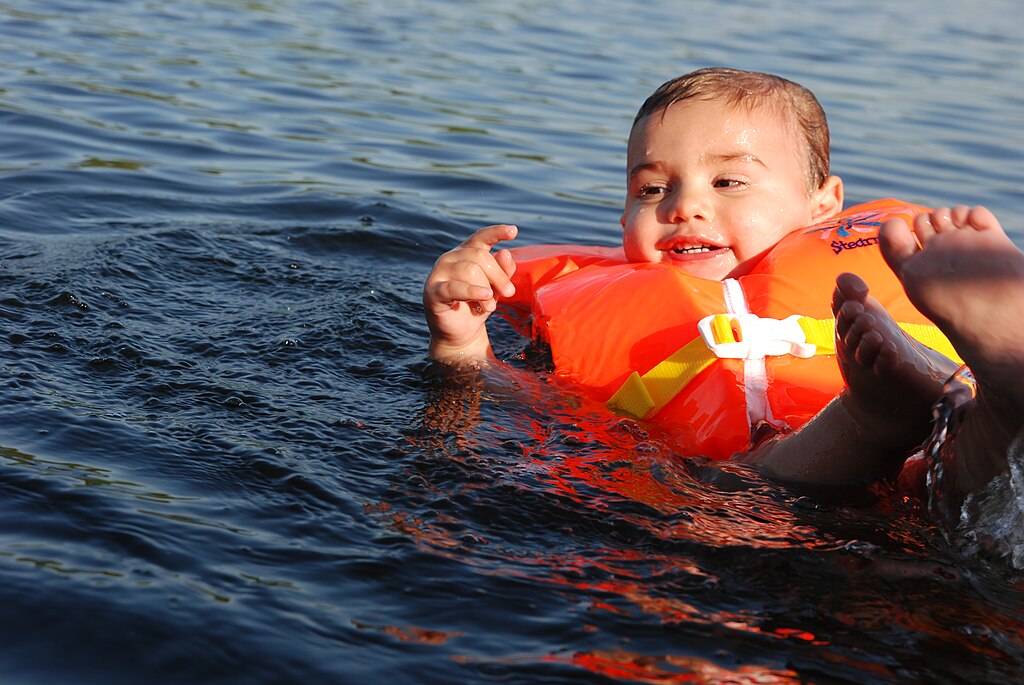 A joyful child wearing an orange life vest floats in calm, sunlit water, smiling and playfully splashing. A pair of adult feet are visible nearby.