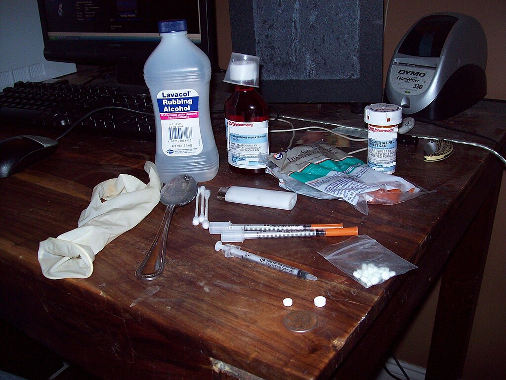 A cluttered wooden desk with medical supplies: a bottle of rubbing alcohol, a latex glove, syringes, medicine bottles, pills, and a spoon, suggesting a medical or clinical setting.