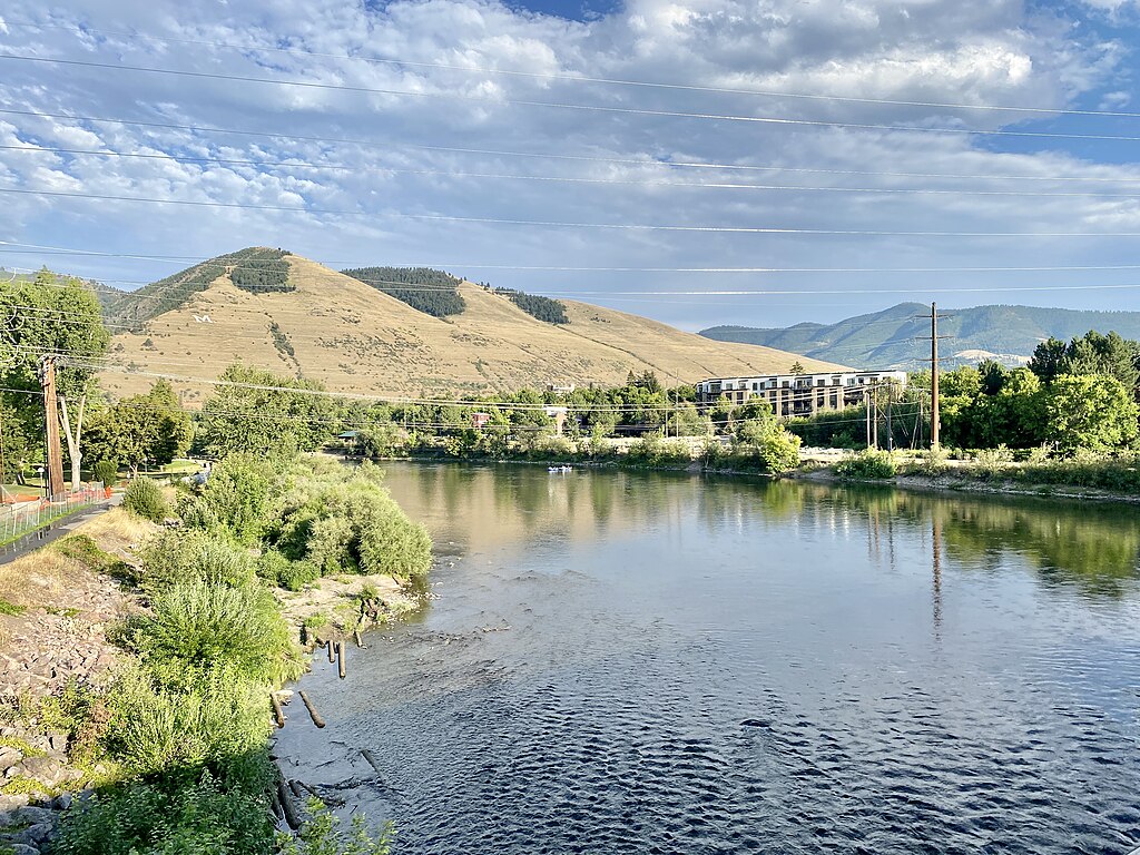 A serene riverside scene under a blue sky, with rolling hills in the background. Lush greenery lines the riverbank, conveying a peaceful, natural setting.