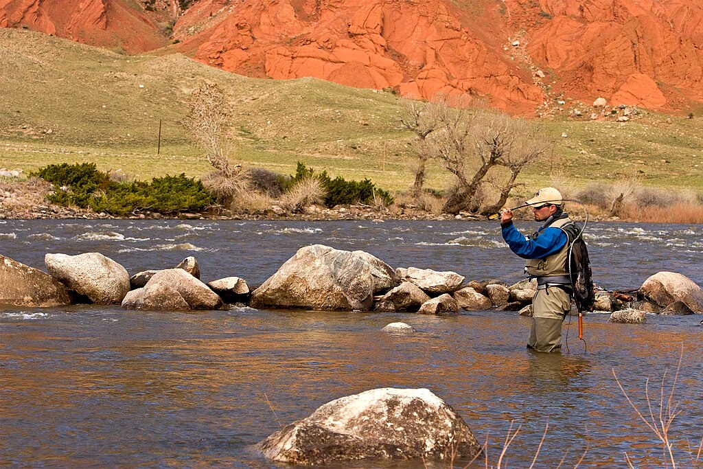 A man fly-fishing in a river, surrounded by rocks, wearing outdoor gear. In the background, vibrant red cliffs and sparse trees create a serene scene.